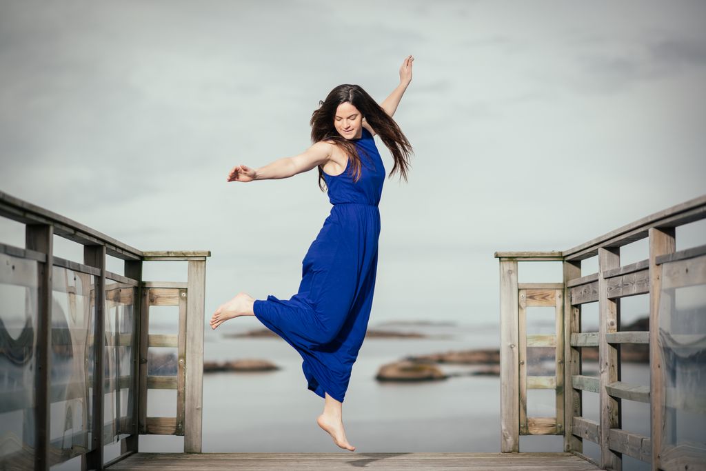 Joyful lifestyle portrait photography featuring woman in blue dress jumping on pier